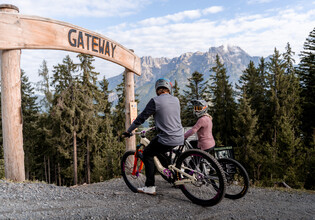Bikepark Einstieg „Gateway“ – Ready to Ride in Leogang | © Yvonne Hörl Zwei Mountainbiker:innen mit Fullface-Helmen stehen mit ihren Bikes unter dem hölzernen „Gateway“-Bogen am Start eines Trails im Bikepark Leogang mit Blick auf das Steinerne Meer. | © Yvonne Hörl