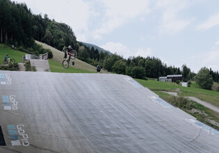 © Yvonne Hörl Ein Biker schiebt sein Rad über den Schotterweg in Richtung Bagjump-Rampe im Übungsareal bei der Talstation Leogang. Rechts ist das große GoPro-Luftkissen für sichere Landungen zu sehen. | © Yvonne Hörl